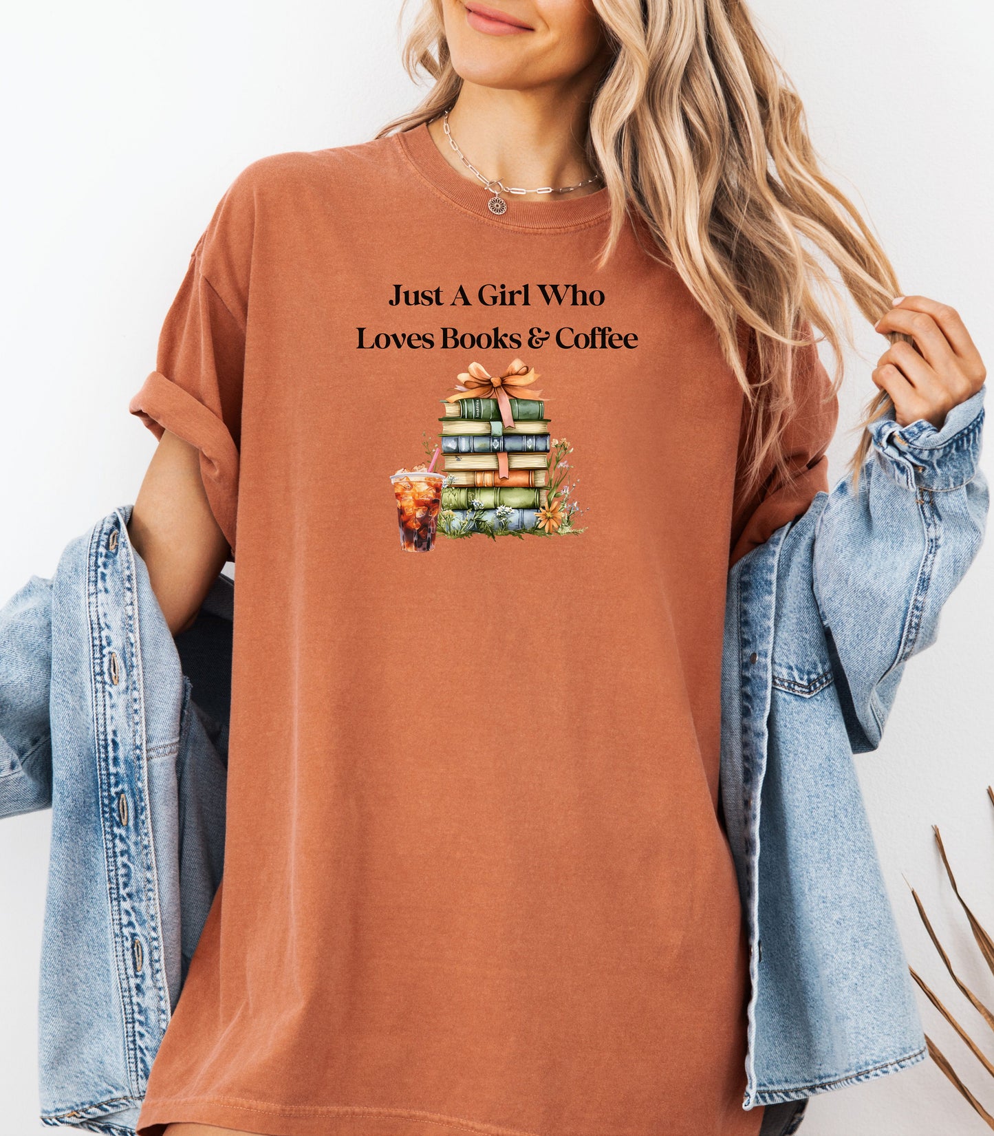Woman wearing a Yam t-shirt with the phrase "Just A Girl Who Loves Books & Coffee" printed above a charming stack of pastel-colored books tied with a ribbon, surrounded by fall foliage and a glass of iced coffee. A cozy and whimsical design perfect for bookworms and coffee lovers alike.