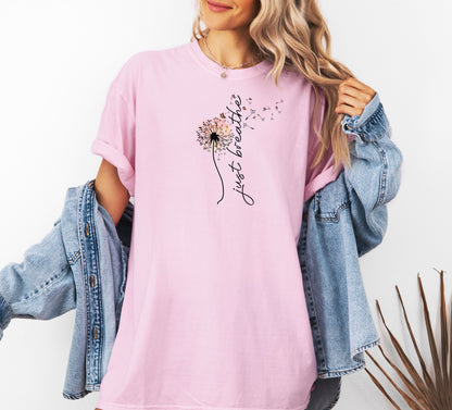 Just Breathe T-shirt featuring an artistic dandelion design with seeds blowing in the wind, transitioning into the handwritten phrase 'just breathe.' Printed on a soft, pastel blossom pink unisex tee, this calming and inspirational graphic is perfect for promoting mindfulness, relaxation, and a peaceful mindset. Ideal for yoga lovers, nature enthusiasts, and casual everyday wear.