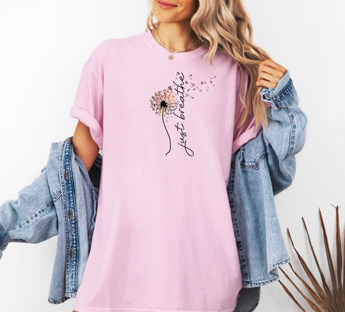 Just Breathe T-shirt featuring an artistic dandelion design with seeds blowing in the wind, transitioning into the handwritten phrase 'just breathe.' Printed on a soft, pastel blossom pink unisex tee, this calming and inspirational graphic is perfect for promoting mindfulness, relaxation, and a peaceful mindset. Ideal for yoga lovers, nature enthusiasts, and casual everyday wear.