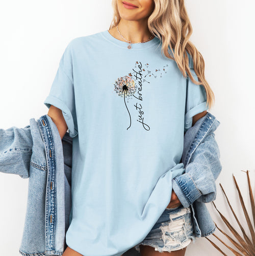 Just Breathe T-shirt featuring an artistic dandelion design with seeds blowing in the wind, transitioning into the handwritten phrase 'just breathe.' Printed on a soft, pastel chambray light blue unisex tee, this calming and inspirational graphic is perfect for promoting mindfulness, relaxation, and a peaceful mindset. Ideal for yoga lovers, nature enthusiasts, and casual everyday wear.
