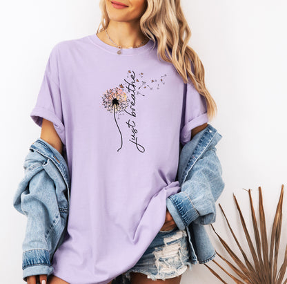 Just Breathe T-shirt featuring an artistic dandelion design with seeds blowing in the wind, transitioning into the handwritten phrase 'just breathe.' Printed on a soft, pastel orchid unisex tee, this calming and inspirational graphic is perfect for promoting mindfulness, relaxation, and a peaceful mindset. Ideal for yoga lovers, nature enthusiasts, and casual everyday wear.
