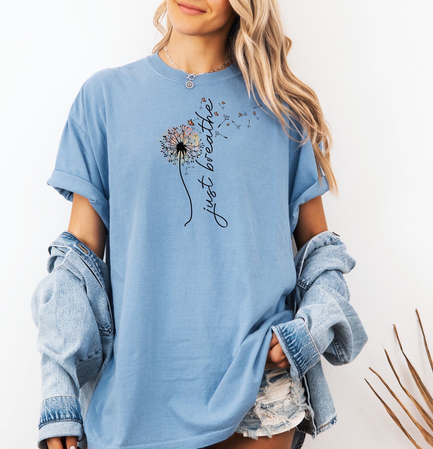 Just Breathe T-shirt featuring an artistic dandelion design with seeds blowing in the wind, transitioning into the handwritten phrase 'just breathe.' Printed on a washed denim unisex tee, this calming and inspirational graphic is perfect for promoting mindfulness, relaxation, and a peaceful mindset. Ideal for yoga lovers, nature enthusiasts, and casual everyday wear.