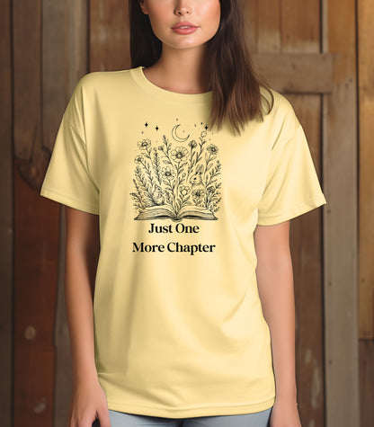 Person wearing a banana soft yellow t-shirt with a charming "Just One More Chapter" design. The graphic features an open book from which wildflowers, stars, and a crescent moon emerge, symbolizing the magic and wonder of getting lost in a good story. Perfect for book lovers and cozy readers.