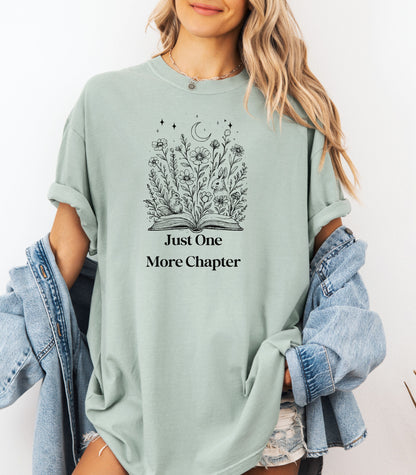 Person wearing a bay light green t-shirt with a charming "Just One More Chapter" design. The graphic features an open book from which wildflowers, stars, and a crescent moon emerge, symbolizing the magic and wonder of getting lost in a good story. Perfect for book lovers and cozy readers.