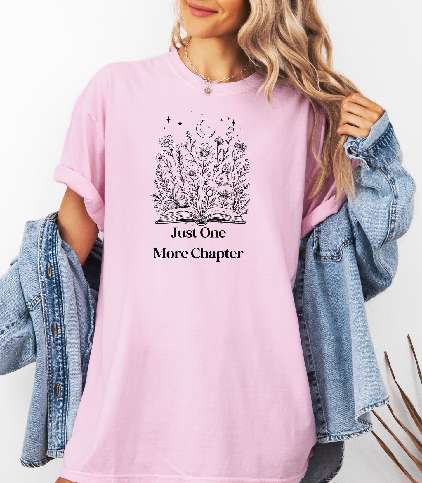 Person wearing a blossom soft pink t-shirt with a charming "Just One More Chapter" design. The graphic features an open book from which wildflowers, stars, and a crescent moon emerge, symbolizing the magic and wonder of getting lost in a good story. Perfect for book lovers and cozy readers.