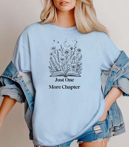 Person wearing a Chambray light blue t-shirt with a charming "Just One More Chapter" design. The graphic features an open book from which wildflowers, stars, and a crescent moon emerge, symbolizing the magic and wonder of getting lost in a good story. Perfect for book lovers and cozy readers.