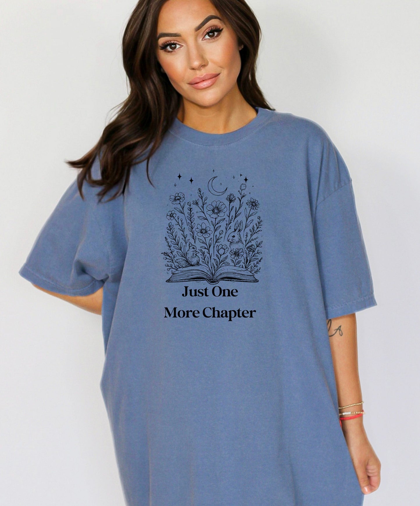 Person wearing a Washed Denim t-shirt with a charming "Just One More Chapter" design. The graphic features an open book from which wildflowers, stars, and a crescent moon emerge, symbolizing the magic and wonder of getting lost in a good story. Perfect for book lovers and cozy readers.