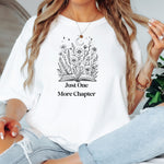 Person wearing a White t-shirt with a charming "Just One More Chapter" design. The graphic features an open book from which wildflowers, stars, and a crescent moon emerge, symbolizing the magic and wonder of getting lost in a good story. Perfect for book lovers and cozy readers.