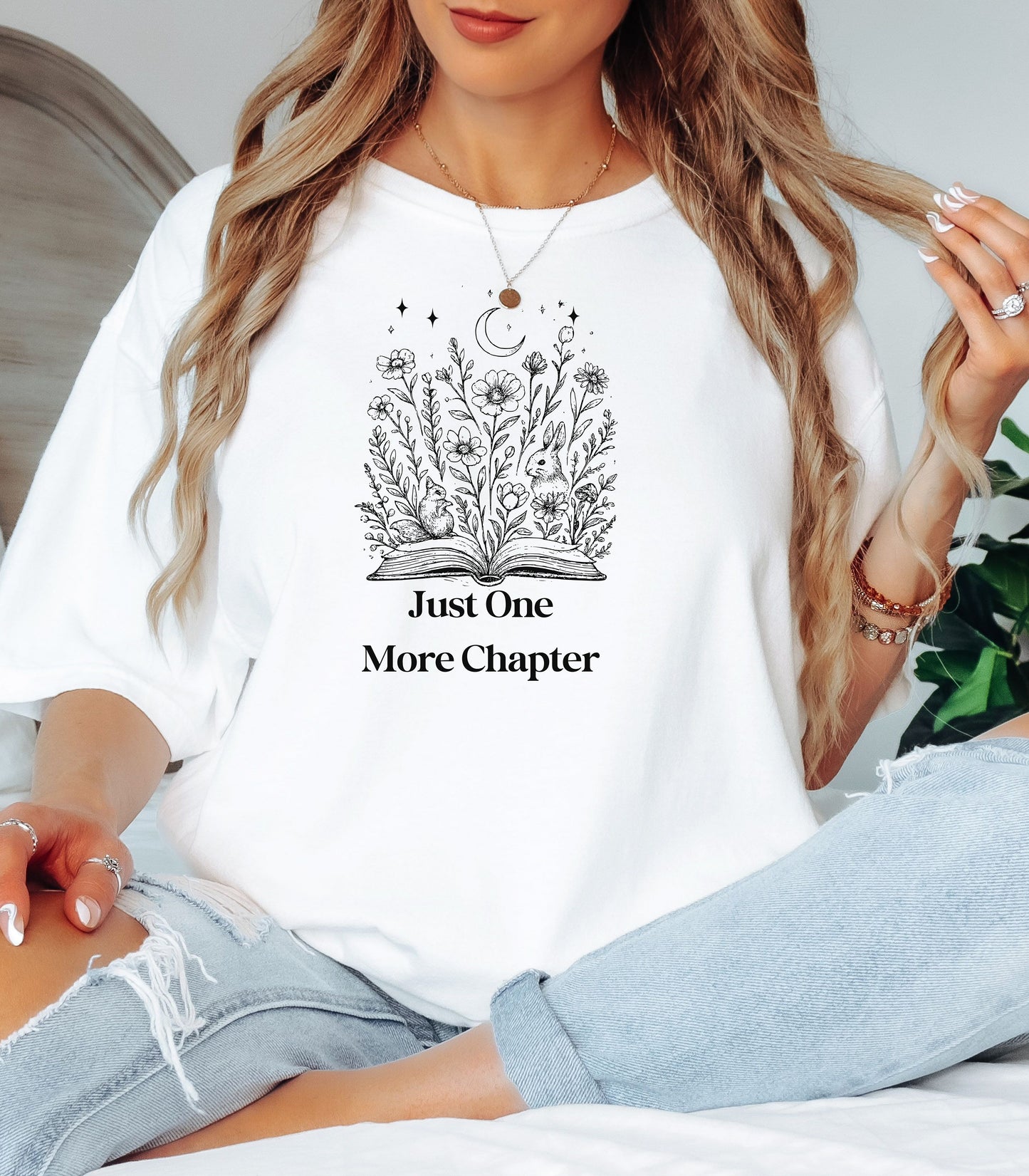 Person wearing a White t-shirt with a charming "Just One More Chapter" design. The graphic features an open book from which wildflowers, stars, and a crescent moon emerge, symbolizing the magic and wonder of getting lost in a good story. Perfect for book lovers and cozy readers.