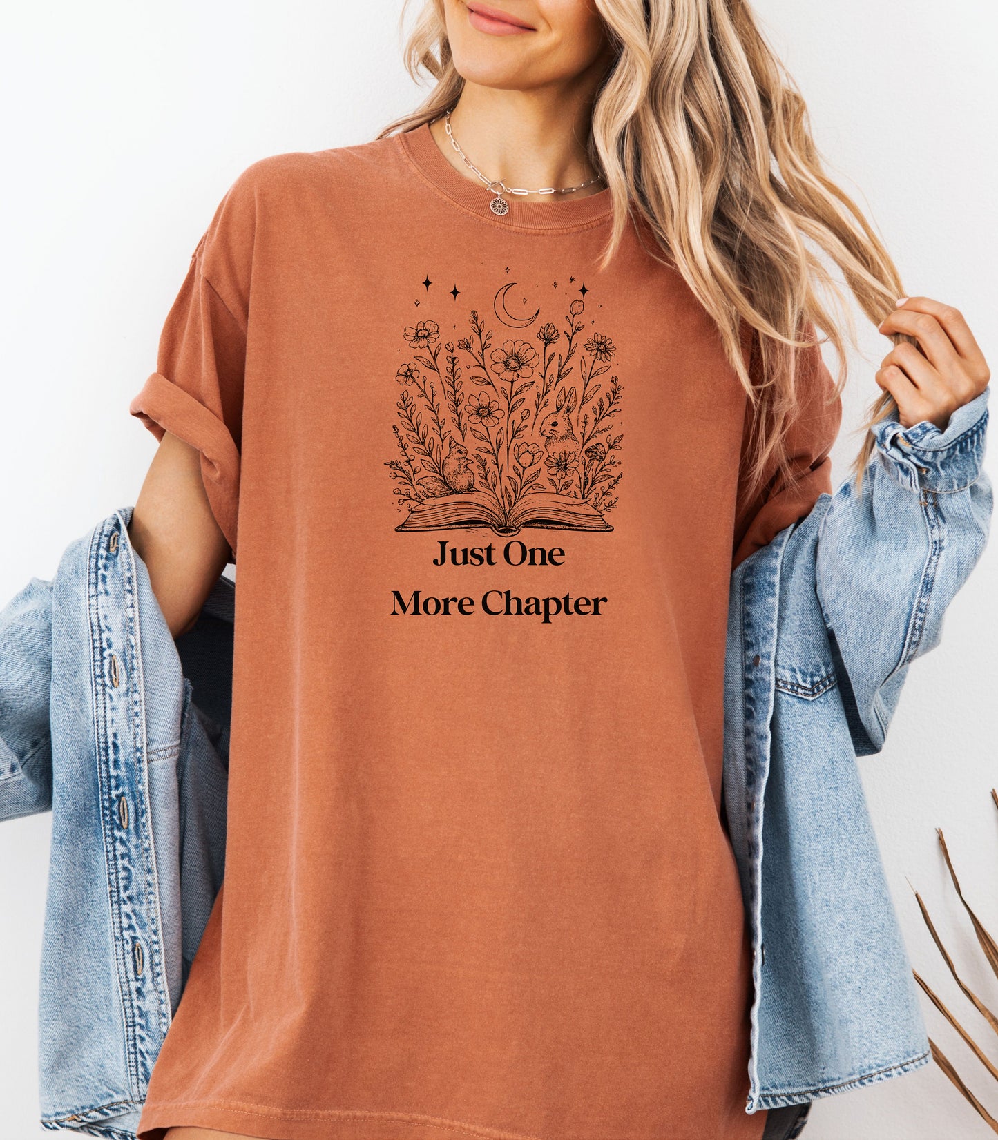 Person wearing a Yam t-shirt with a charming "Just One More Chapter" design. The graphic features an open book from which wildflowers, stars, and a crescent moon emerge, symbolizing the magic and wonder of getting lost in a good story. Perfect for book lovers and cozy readers.