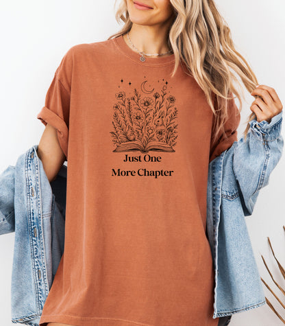 Person wearing a Yam t-shirt with a charming "Just One More Chapter" design. The graphic features an open book from which wildflowers, stars, and a crescent moon emerge, symbolizing the magic and wonder of getting lost in a good story. Perfect for book lovers and cozy readers.