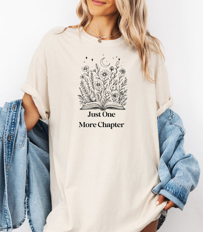 Person wearing an Ivory t-shirt with a charming "Just One More Chapter" design. The graphic features an open book from which wildflowers, stars, and a crescent moon emerge, symbolizing the magic and wonder of getting lost in a good story. Perfect for book lovers and cozy readers.
