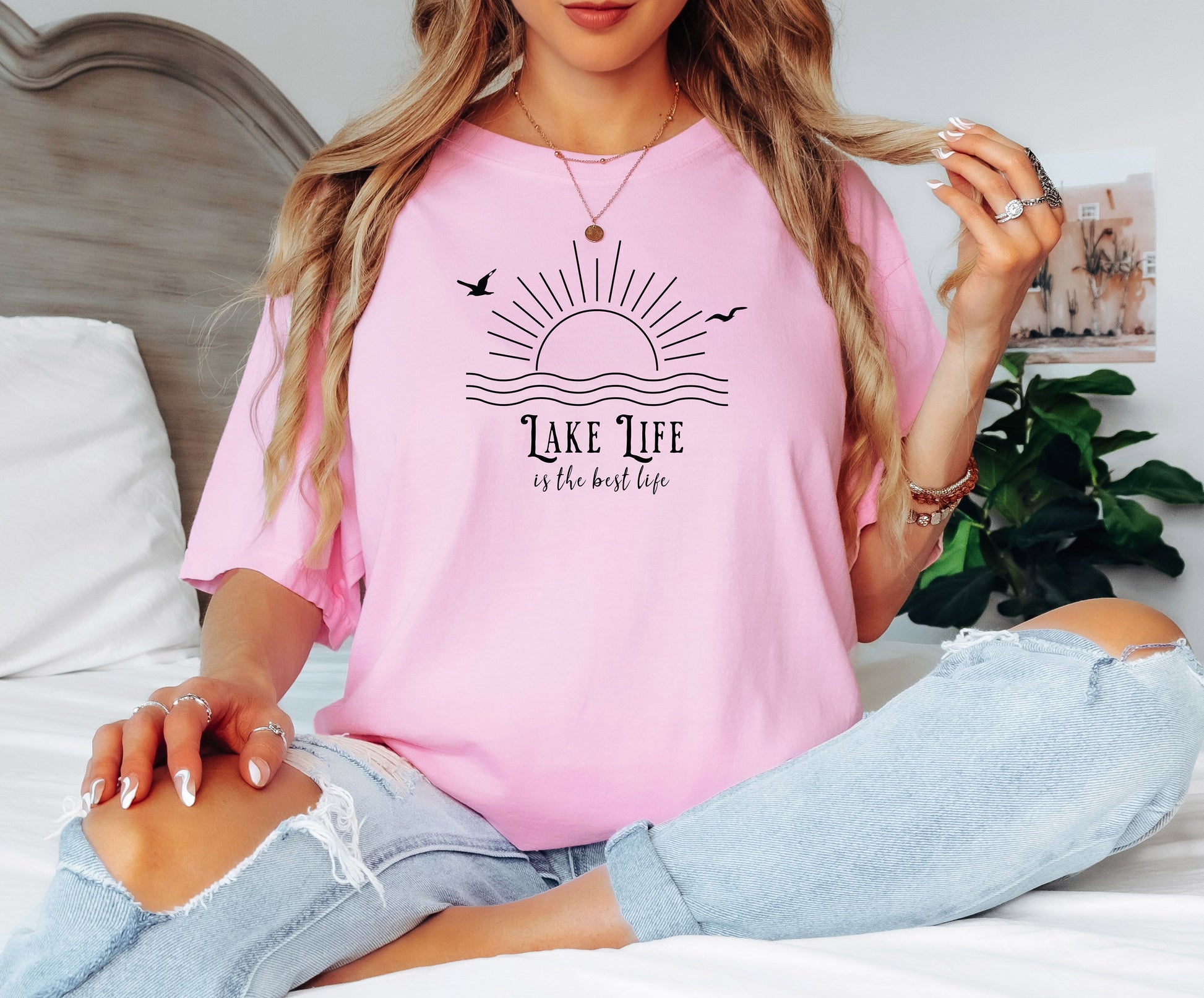 Person wearing a blossom pink T-shirt featuring a minimalist black line art design of a rising sun over wavy water, with two flying birds. The text reads “Lake Life is the best life” in elegant, mixed fonts. A relaxed, nature-inspired design perfect for lake lovers, summer days, and casual outdoor adventures.