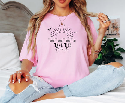 Person wearing a blossom pink T-shirt featuring a minimalist black line art design of a rising sun over wavy water, with two flying birds. The text reads “Lake Life is the best life” in elegant, mixed fonts. A relaxed, nature-inspired design perfect for lake lovers, summer days, and casual outdoor adventures.