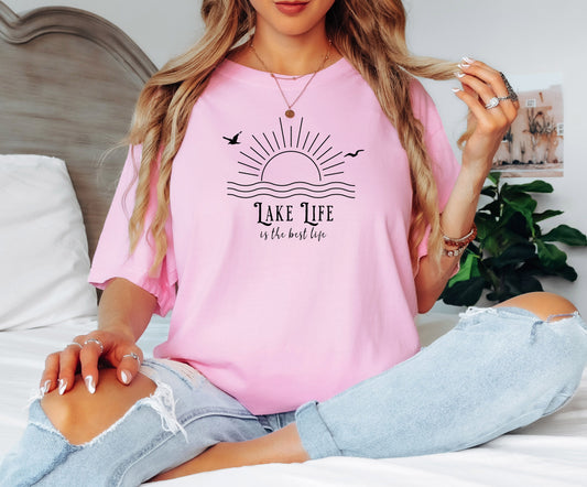 Person wearing a blossom pink T-shirt featuring a minimalist black line art design of a rising sun over wavy water, with two flying birds. The text reads “Lake Life is the best life” in elegant, mixed fonts. A relaxed, nature-inspired design perfect for lake lovers, summer days, and casual outdoor adventures.
