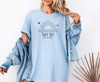 Person wearing a chambray light blue T-shirt featuring a minimalist black line art design of a rising sun over wavy water, with two flying birds. The text reads “Lake Life is the best life” in elegant, mixed fonts. A relaxed, nature-inspired design perfect for lake lovers, summer days, and casual outdoor adventures.