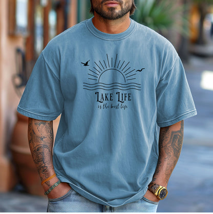 Person wearing a washed denim T-shirt featuring a minimalist black line art design of a rising sun over wavy water, with two flying birds. The text reads “Lake Life is the best life” in elegant, mixed fonts. A relaxed, nature-inspired design perfect for lake lovers, summer days, and casual outdoor adventures.