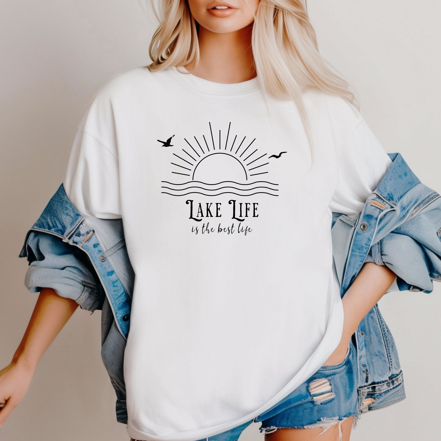 Person wearing a classic white T-shirt featuring a minimalist black line art design of a rising sun over wavy water, with two flying birds. The text reads “Lake Life is the best life” in elegant, mixed fonts. A relaxed, nature-inspired design perfect for lake lovers, summer days, and casual outdoor adventures.