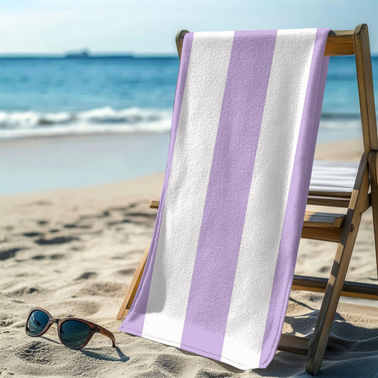 A bold vertical lavender and white striped beach towel hanging over a beach chair with the ocean behind it. The fresh, classic striped design brings a soft and stylish touch to any seaside escape, perfect for lounging, drying off, or making a beachside statement.