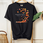 Black Comfort Colors T-shirt featuring a fall-themed design with swirling autumn leaves and the phrase "Leaves Are Falling, Autumn is Calling" in elegant white script. The vibrant orange, red, and yellow leaves create a whimsical motion effect. Shirt is styled with a front knot and displayed on a wooden hanger in a cozy, natural setting.