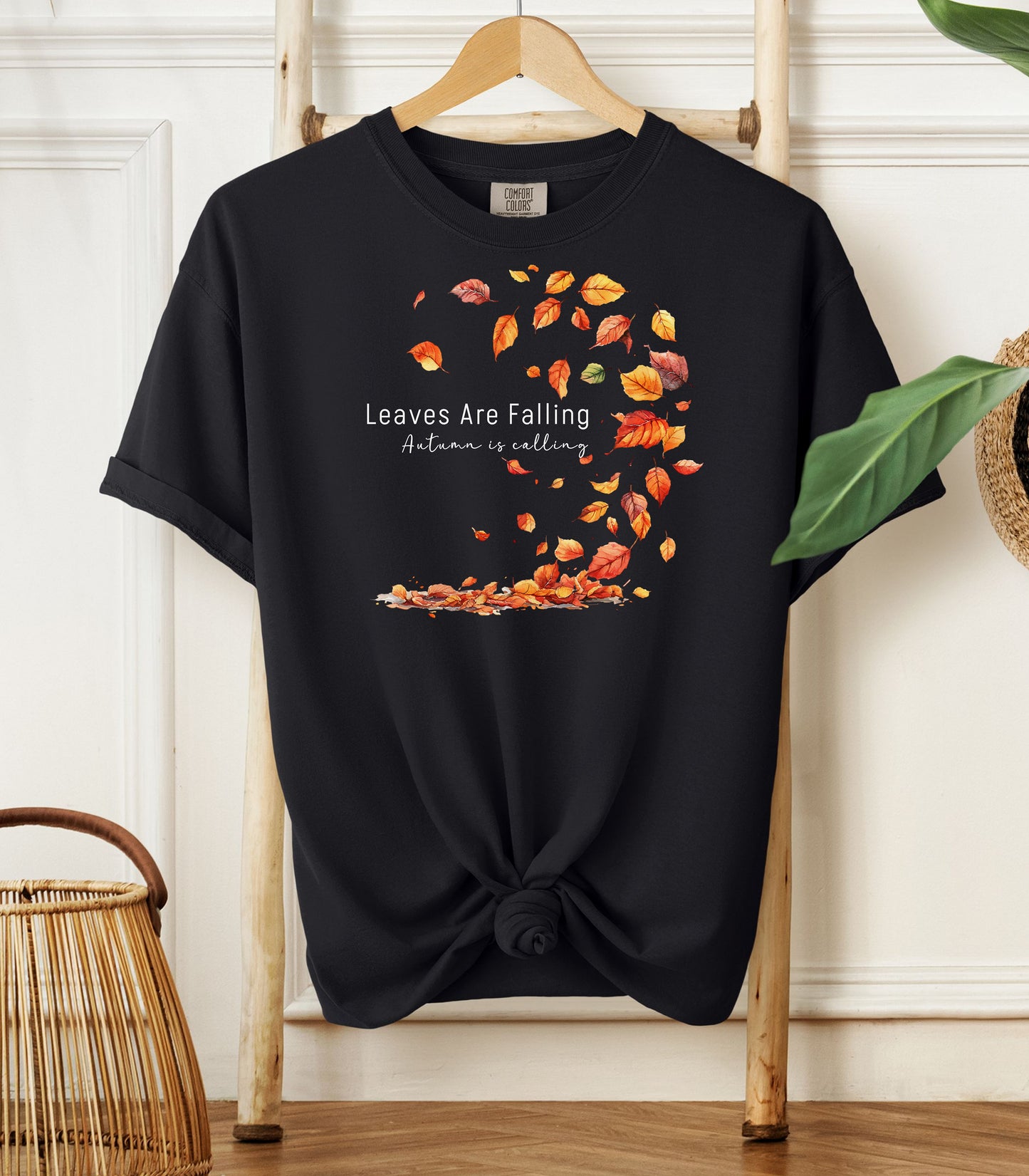 Black Comfort Colors T-shirt featuring a fall-themed design with swirling autumn leaves and the phrase "Leaves Are Falling, Autumn is Calling" in elegant white script. The vibrant orange, red, and yellow leaves create a whimsical motion effect. Shirt is styled with a front knot and displayed on a wooden hanger in a cozy, natural setting.