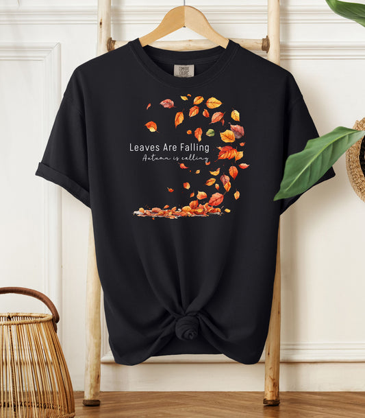 Black Comfort Colors T-shirt featuring a fall-themed design with swirling autumn leaves and the phrase "Leaves Are Falling, Autumn is Calling" in elegant white script. The vibrant orange, red, and yellow leaves create a whimsical motion effect. Shirt is styled with a front knot and displayed on a wooden hanger in a cozy, natural setting.