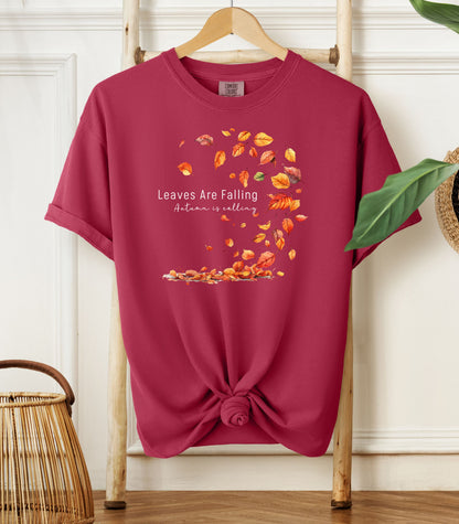 Chili Comfort Colors T-shirt featuring a fall-themed design with swirling autumn leaves and the phrase "Leaves Are Falling, Autumn is Calling" in elegant white script. The vibrant orange, red, and yellow leaves create a whimsical motion effect. Shirt is styled with a front knot and displayed on a wooden hanger in a cozy, natural setting.