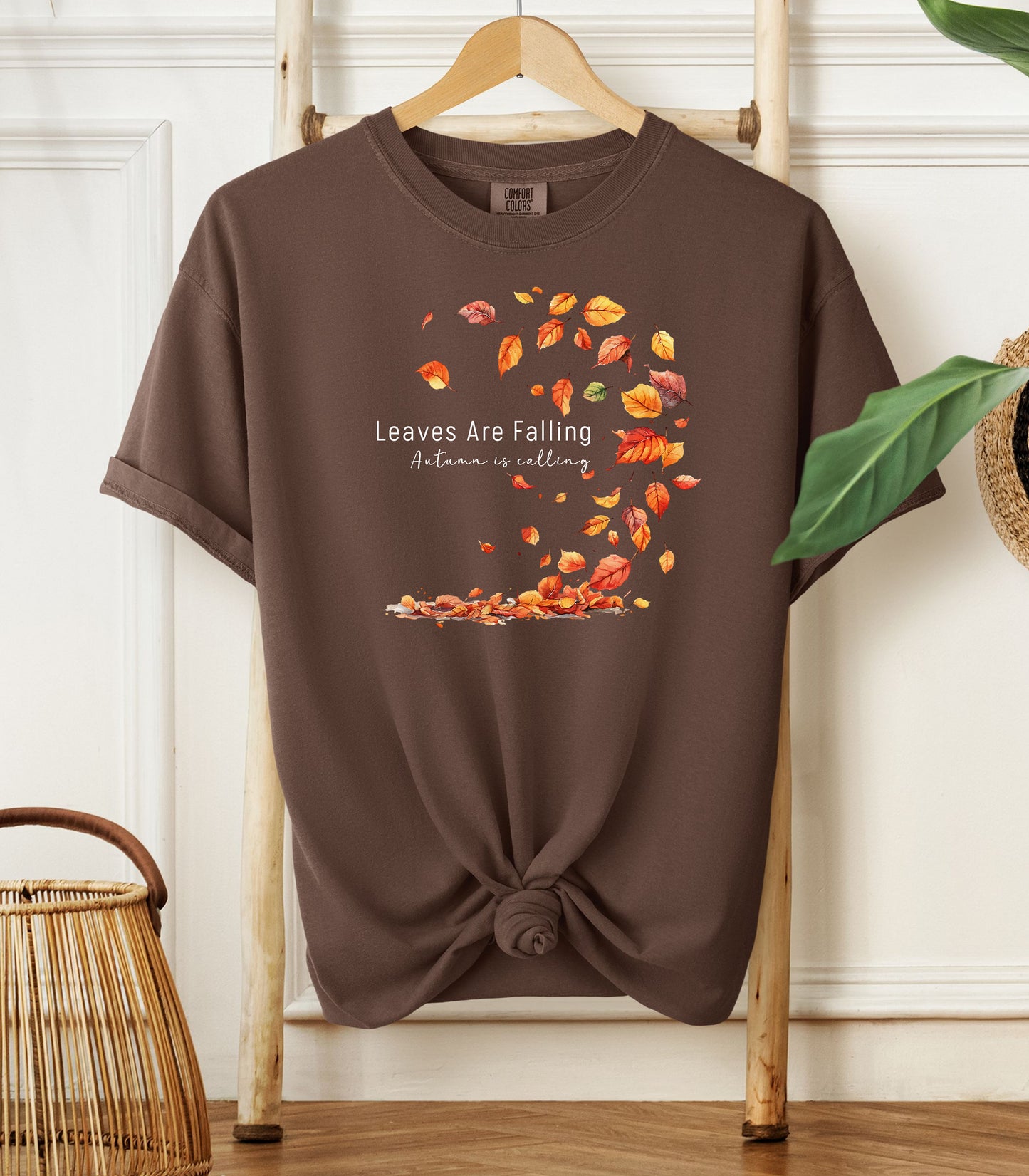 Espresso Comfort Colors T-shirt featuring a fall-themed design with swirling autumn leaves and the phrase "Leaves Are Falling, Autumn is Calling" in elegant white script. The vibrant orange, red, and yellow leaves create a whimsical motion effect. Shirt is styled with a front knot and displayed on a wooden hanger in a cozy, natural setting.