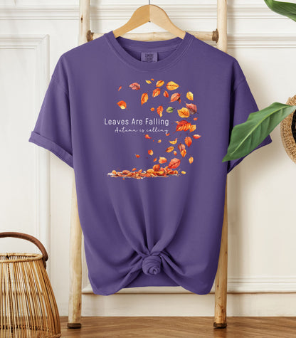 Grape Comfort Colors T-shirt featuring a fall-themed design with swirling autumn leaves and the phrase "Leaves Are Falling, Autumn is Calling" in elegant white script. The vibrant orange, red, and yellow leaves create a whimsical motion effect. Shirt is styled with a front knot and displayed on a wooden hanger in a cozy, natural setting.