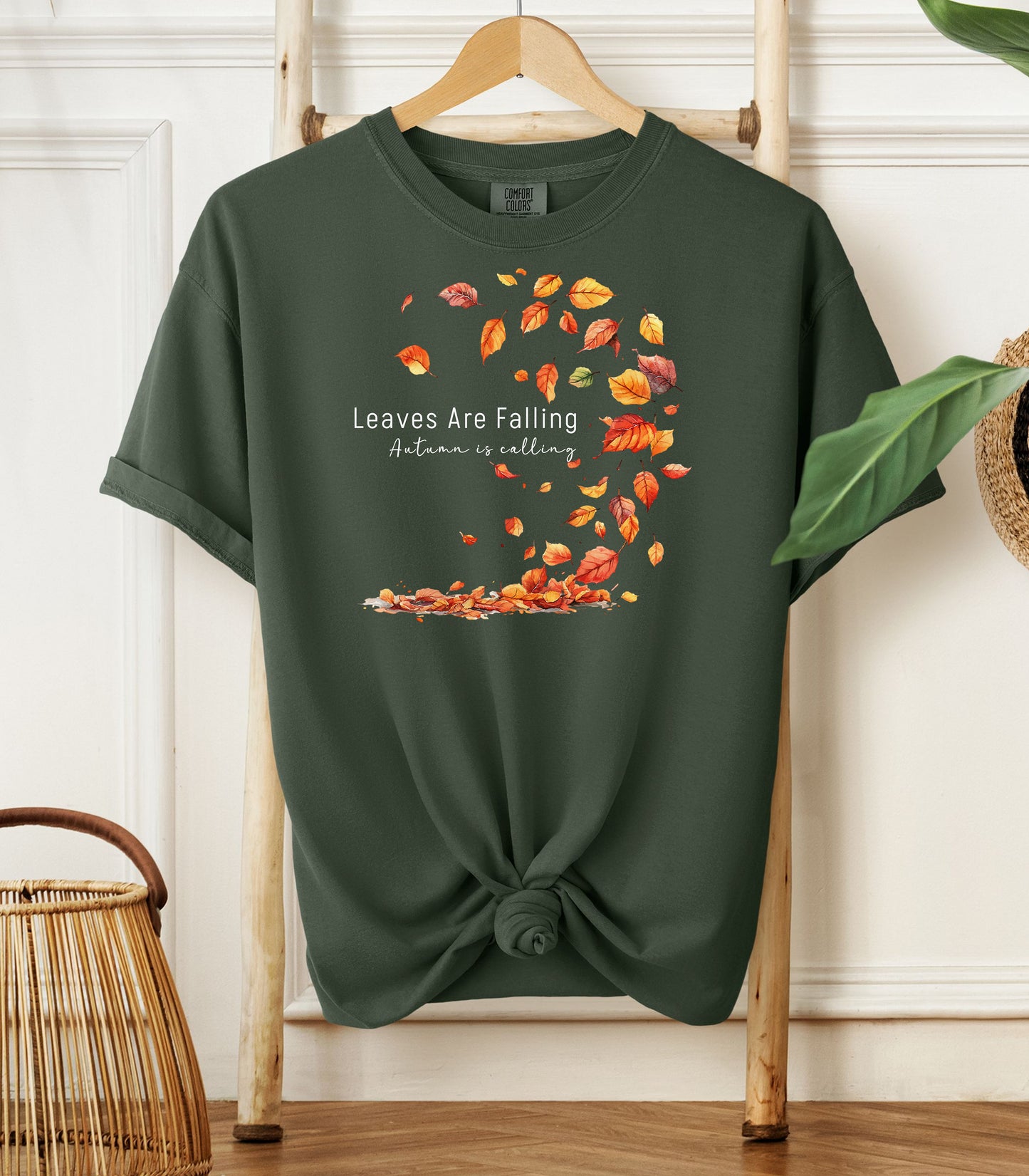 Hemp color Comfort Colors T-shirt featuring a fall-themed design with swirling autumn leaves and the phrase "Leaves Are Falling, Autumn is Calling" in elegant white script. The vibrant orange, red, and yellow leaves create a whimsical motion effect. Shirt is styled with a front knot and displayed on a wooden hanger in a cozy, natural setting.