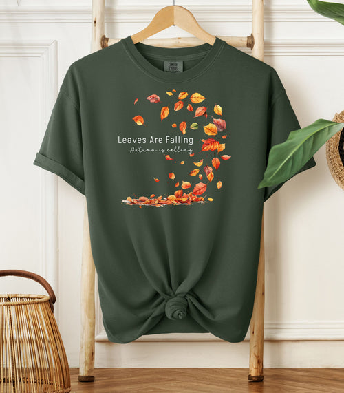Hemp color Comfort Colors T-shirt featuring a fall-themed design with swirling autumn leaves and the phrase "Leaves Are Falling, Autumn is Calling" in elegant white script. The vibrant orange, red, and yellow leaves create a whimsical motion effect. Shirt is styled with a front knot and displayed on a wooden hanger in a cozy, natural setting.