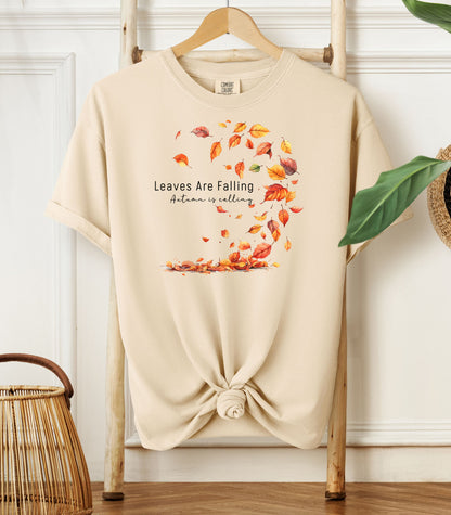 Ivory Comfort Colors T-shirt featuring a fall-themed design with swirling autumn leaves and the phrase "Leaves Are Falling, Autumn is Calling" in elegant black script. The vibrant orange, red, and yellow leaves create a whimsical motion effect. Shirt is styled with a front knot and displayed on a wooden hanger in a cozy, natural setting.