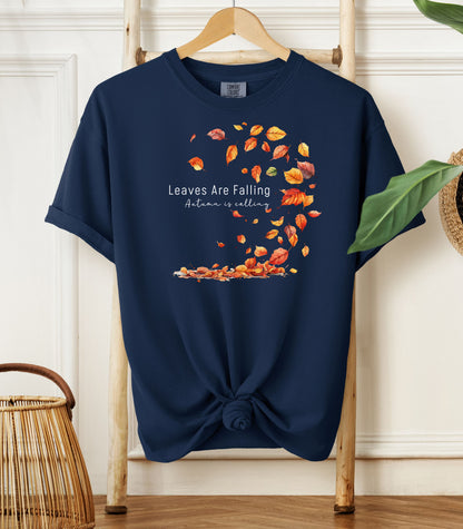 True Navy Comfort Colors T-shirt featuring a fall-themed design with swirling autumn leaves and the phrase "Leaves Are Falling, Autumn is Calling" in elegant white script. The vibrant orange, red, and yellow leaves create a whimsical motion effect. Shirt is styled with a front knot and displayed on a wooden hanger in a cozy, natural setting.