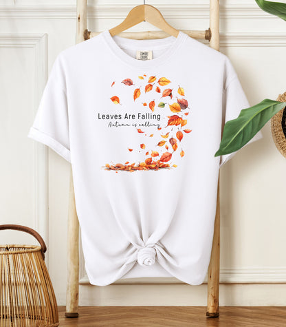 White Comfort Colors T-shirt featuring a fall-themed design with swirling autumn leaves and the phrase "Leaves Are Falling, Autumn is Calling" in elegant black script. The vibrant orange, red, and yellow leaves create a whimsical motion effect. Shirt is styled with a front knot and displayed on a wooden hanger in a cozy, natural setting.