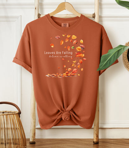 Yam Comfort Colors T-shirt featuring a fall-themed design with swirling autumn leaves and the phrase "Leaves Are Falling, Autumn is Calling" in elegant white script. The vibrant orange, red, and yellow leaves create a whimsical motion effect. Shirt is styled with a front knot and displayed on a wooden hanger in a cozy, natural setting.