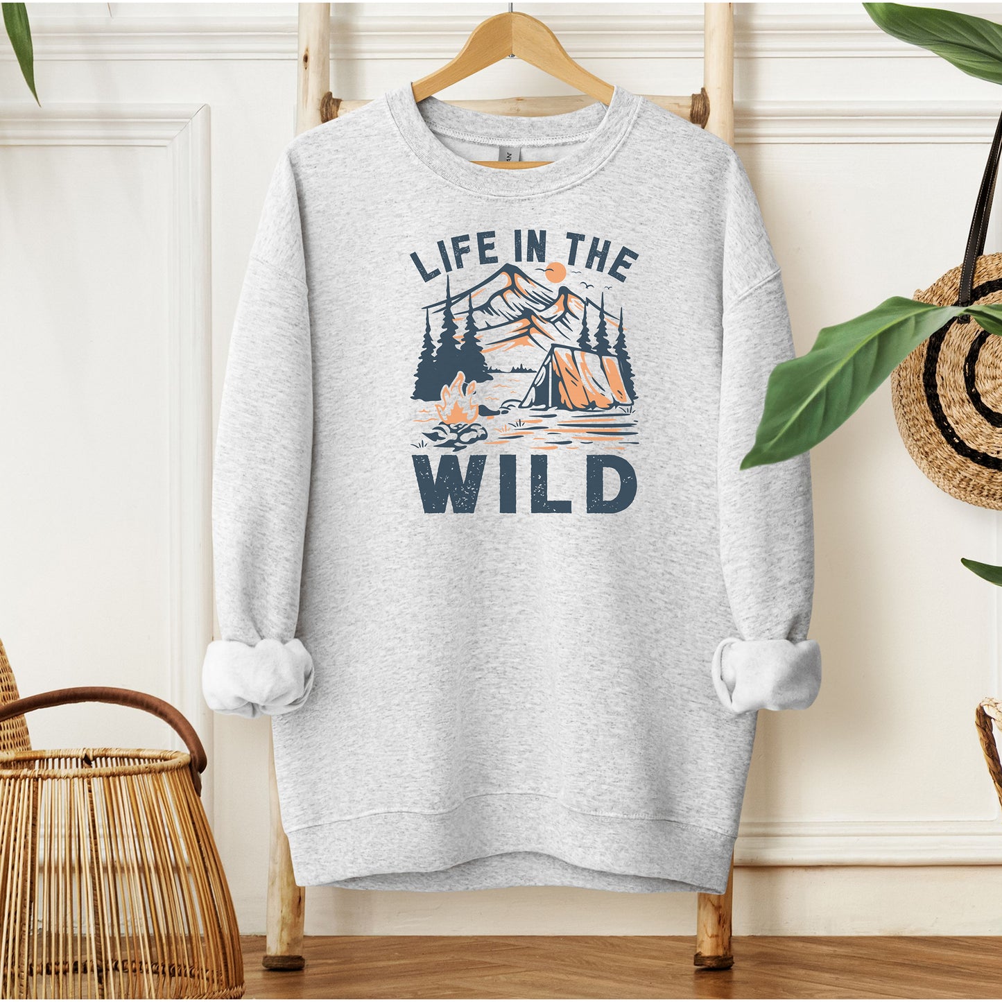 Ash sweatshirt featuring the “Life in the Wild” design with a vintage-style outdoor scene of a tent, campfire, and pine trees set against a mountain backdrop and sun. The rustic illustration in muted orange and deep gray captures the spirit of camping, adventure, and nature lovers. Perfect for hikers, outdoor enthusiasts, and those who embrace the wild. Cozy, casual style ideal for layering on cool days in the great outdoors or relaxing at home.