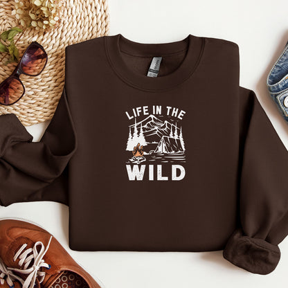 "Life In The Wild graphic on a dark chocolate sweatshirt, featuring a white illustration of a mountain range, pine trees, a tent, and a campfire with orange flames. The design has a rustic, outdoorsy feel, perfect for camping and nature lovers. Ideal cozy apparel for adventurers, hikers, and outdoor enthusiasts seeking a rugged yet stylish look."