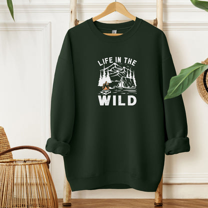 "Life In The Wild graphic on a forest green sweatshirt, featuring a white illustration of a mountain range, pine trees, a tent, and a campfire with orange flames. The design has a rustic, outdoorsy feel, perfect for camping and nature lovers. Ideal cozy apparel for adventurers, hikers, and outdoor enthusiasts seeking a rugged yet stylish look."