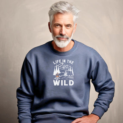 "Life In The Wild graphic on a heather sport dark navy sweatshirt, featuring a white illustration of a mountain range, pine trees, a tent, and a campfire with orange flames. The design has a rustic, outdoorsy feel, perfect for camping and nature lovers. Ideal cozy apparel for adventurers, hikers, and outdoor enthusiasts seeking a rugged yet stylish look."