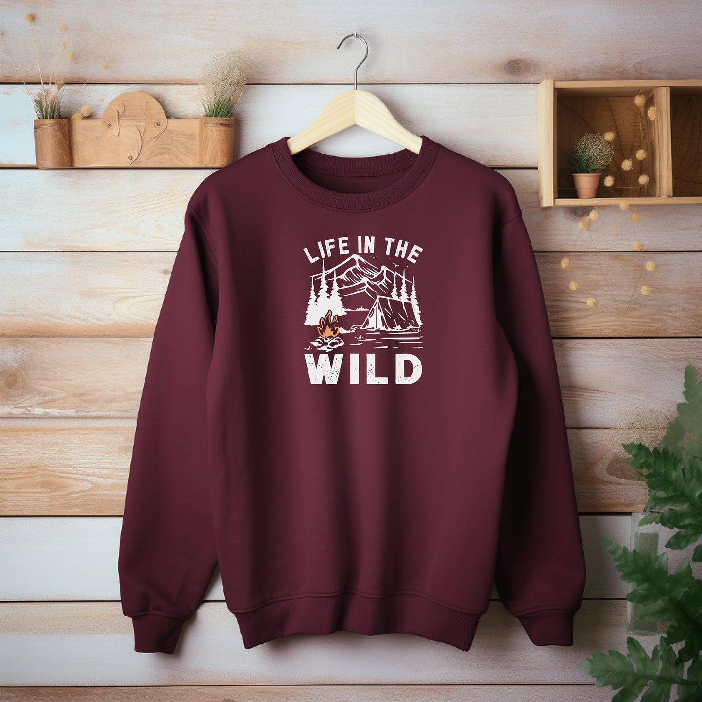 "Life In The Wild graphic on a maroon sweatshirt, featuring a white illustration of a mountain range, pine trees, a tent, and a campfire with orange flames. The design has a rustic, outdoorsy feel, perfect for camping and nature lovers. Ideal cozy apparel for adventurers, hikers, and outdoor enthusiasts seeking a rugged yet stylish look."