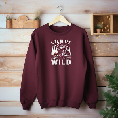 "Life In The Wild graphic on a maroon sweatshirt, featuring a white illustration of a mountain range, pine trees, a tent, and a campfire with orange flames. The design has a rustic, outdoorsy feel, perfect for camping and nature lovers. Ideal cozy apparel for adventurers, hikers, and outdoor enthusiasts seeking a rugged yet stylish look."