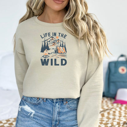 Sand sweatshirt featuring the “Life in the Wild” design with a vintage-style outdoor scene of a tent, campfire, and pine trees set against a mountain backdrop and sun. The rustic illustration in muted orange and deep gray captures the spirit of camping, adventure, and nature lovers. Perfect for hikers, outdoor enthusiasts, and those who embrace the wild. Cozy, casual style ideal for layering on cool days in the great outdoors or relaxing at home.