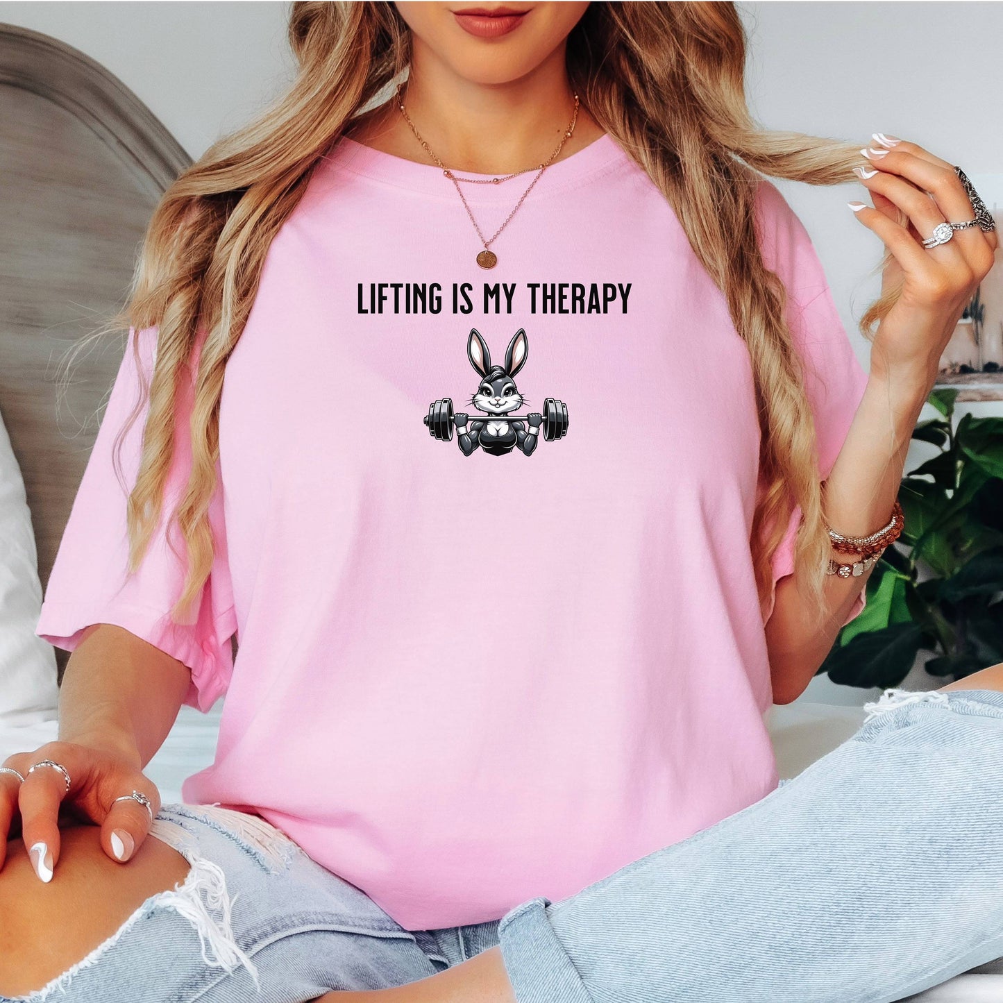 Person wearing a blossom pink T-shirt featuring the phrase “Lifting Is My Therapy” above a muscular cartoon rabbit holding a barbell with heavy weights. The playful yet powerful design blends fitness motivation with humor, making it perfect for weightlifters, gym-goers, and strength training enthusiasts who find stress relief in lifting.