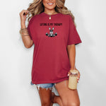 Person wearing a chili red T-shirt featuring the phrase “Lifting Is My Therapy” above a muscular cartoon rabbit holding a barbell with heavy weights. The playful yet powerful design blends fitness motivation with humor, making it perfect for weightlifters, gym-goers, and strength training enthusiasts who find stress relief in lifting.