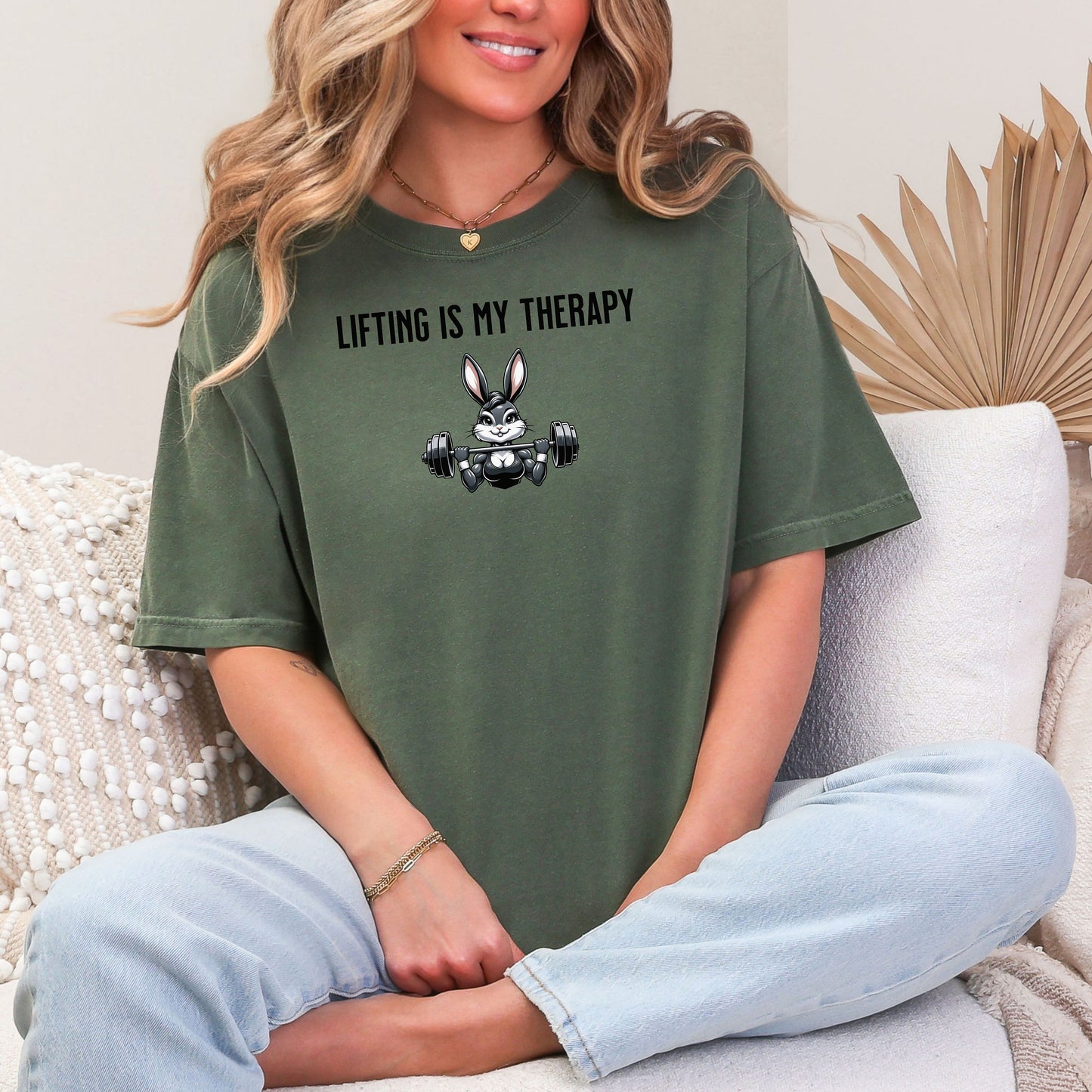 Person wearing a hemp color T-shirt featuring the phrase “Lifting Is My Therapy” above a muscular cartoon rabbit holding a barbell with heavy weights. The playful yet powerful design blends fitness motivation with humor, making it perfect for weightlifters, gym-goers, and strength training enthusiasts who find stress relief in lifting.