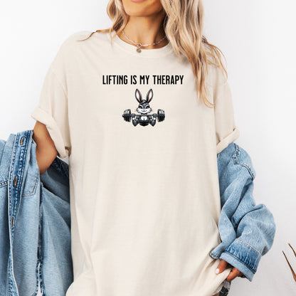 Person wearing an ivory T-shirt featuring the phrase “Lifting Is My Therapy” above a muscular cartoon rabbit holding a barbell with heavy weights. The playful yet powerful design blends fitness motivation with humor, making it perfect for weightlifters, gym-goers, and strength training enthusiasts who find stress relief in lifting.
