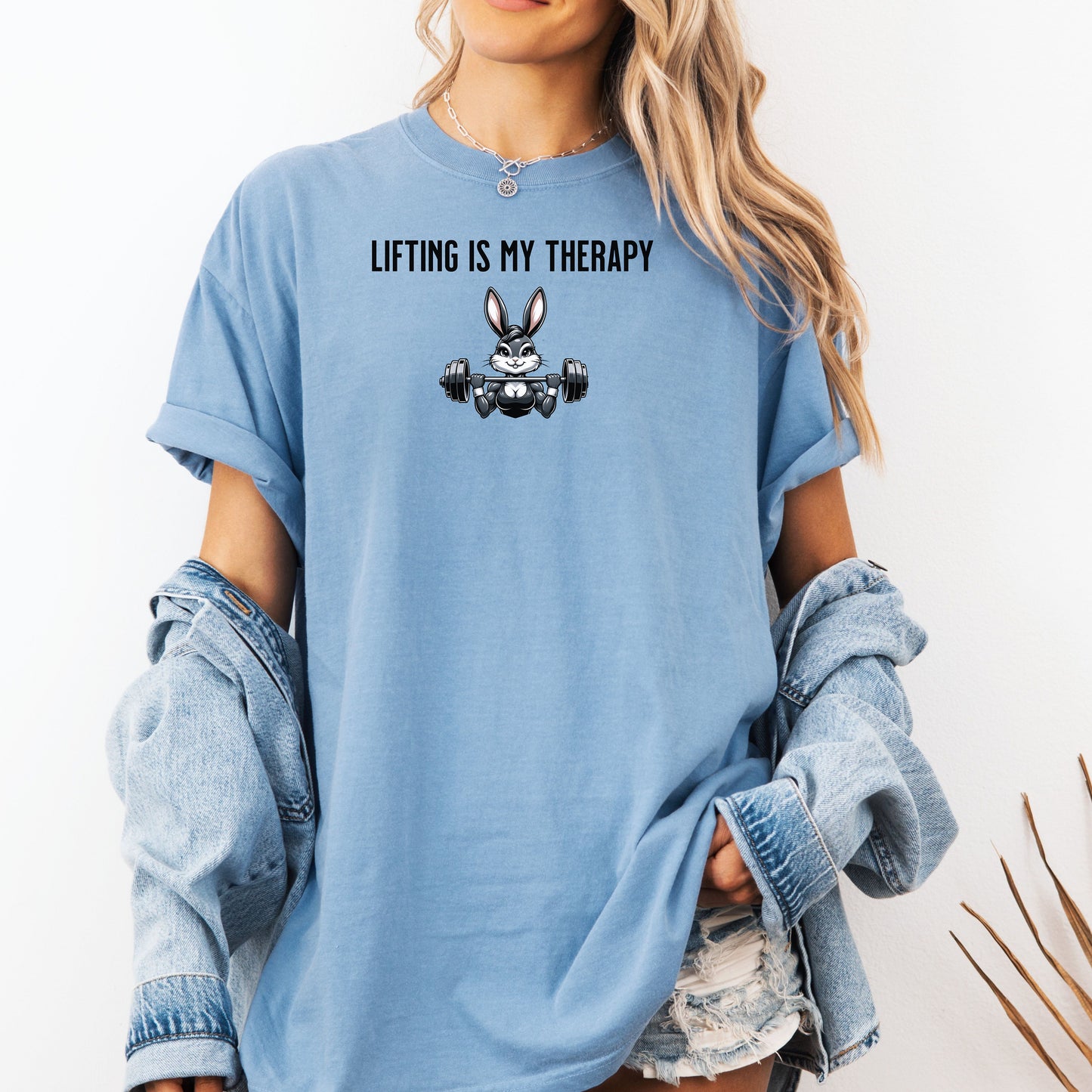 Person wearing a washed denim T-shirt featuring the phrase “Lifting Is My Therapy” above a muscular cartoon rabbit holding a barbell with heavy weights. The playful yet powerful design blends fitness motivation with humor, making it perfect for weightlifters, gym-goers, and strength training enthusiasts who find stress relief in lifting.