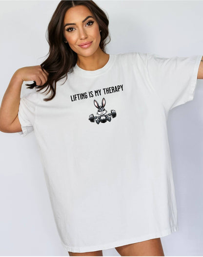 Person wearing a classic white T-shirt featuring the phrase “Lifting Is My Therapy” above a muscular cartoon rabbit holding a barbell with heavy weights. The playful yet powerful design blends fitness motivation with humor, making it perfect for weightlifters, gym-goers, and strength training enthusiasts who find stress relief in lifting.