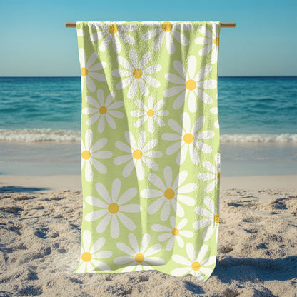 Light green beach towel hanging down from a stand featuring a cheerful retro daisy pattern with large white petals and yellow centers. Set on a sunny beach with ocean waves in the background and sunglasses in the sand, this towel brings playful vintage vibes to any seaside or poolside day.
