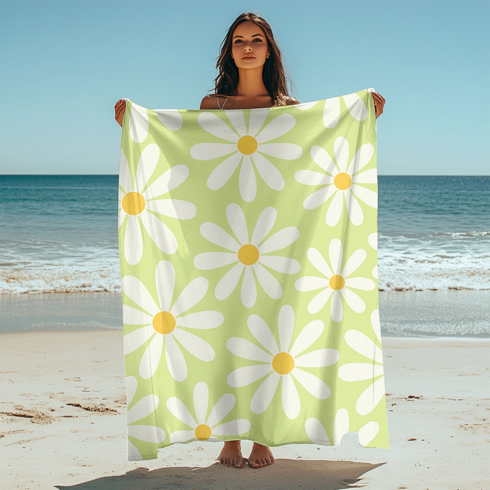 Woman holding up a light green beach towel, featuring a cheerful retro daisy pattern with large white petals and yellow centers. Set on a sunny beach with ocean waves in the background and sunglasses in the sand, this towel brings playful vintage vibes to any seaside or poolside day.